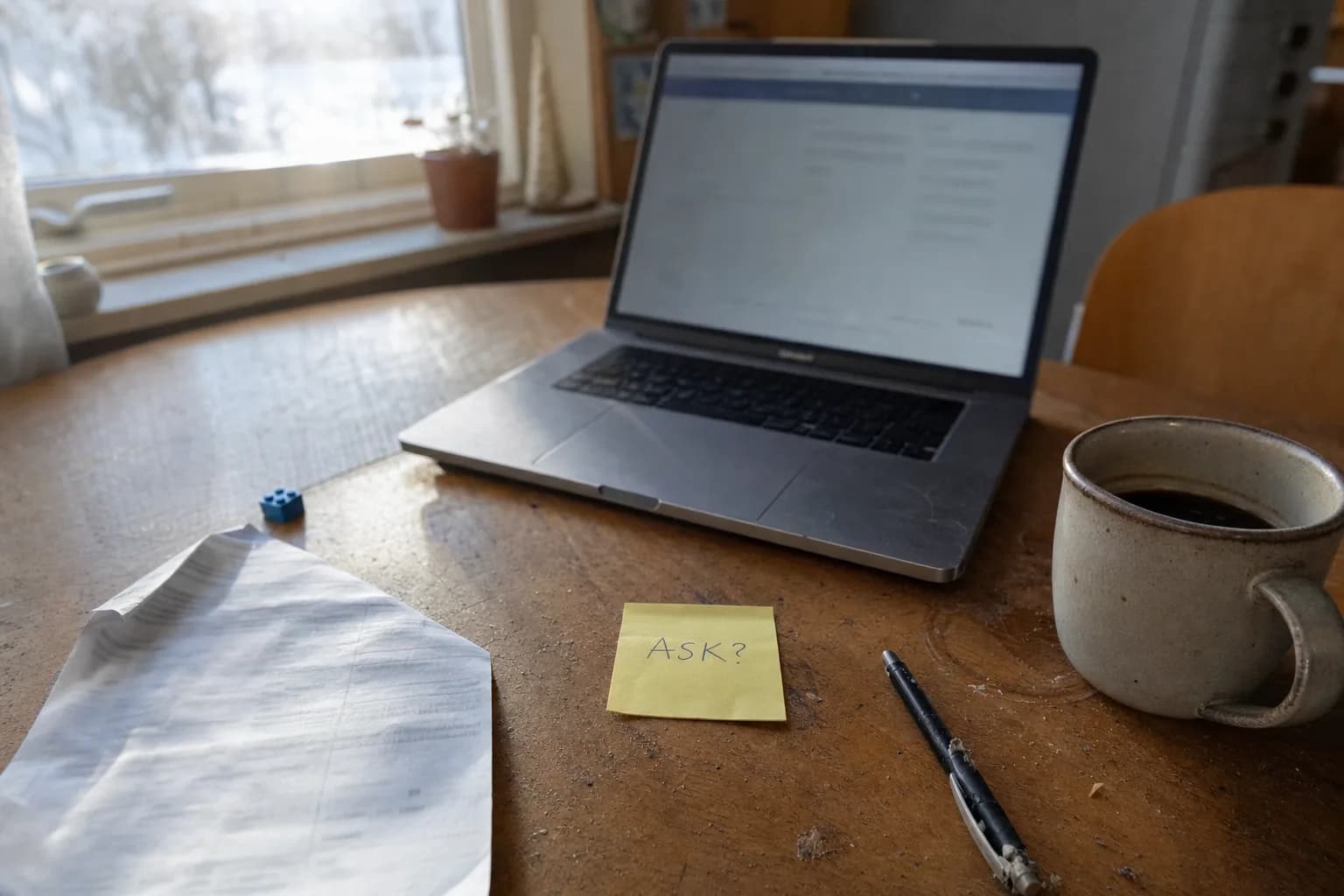 Kjøkkenbord med laptop, kaffekopp og en gul lapp med "ASK?" skrevet på